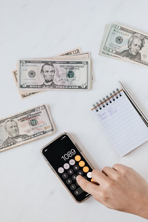 A hand using a smartphone calculator, surrounded by dollar bills and a notepad with financial notes, illustrating financial planning and management.
