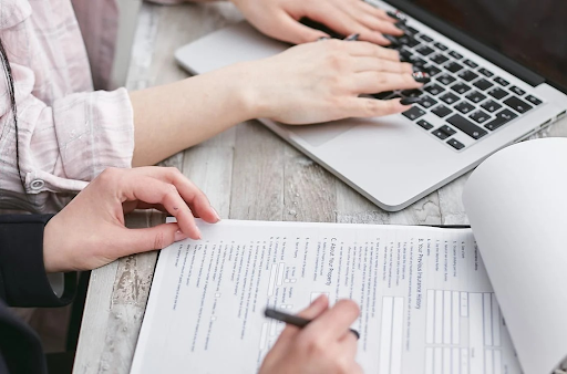 Two people working together, one typing on a laptop and the other filling out forms, indicating collaboration in financial planning.