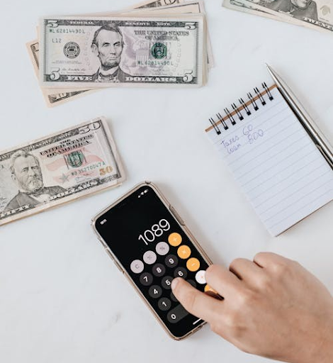 A person using a calculator with U.S. dollar bills and a notebook nearby, emphasizing debt calculation and management.