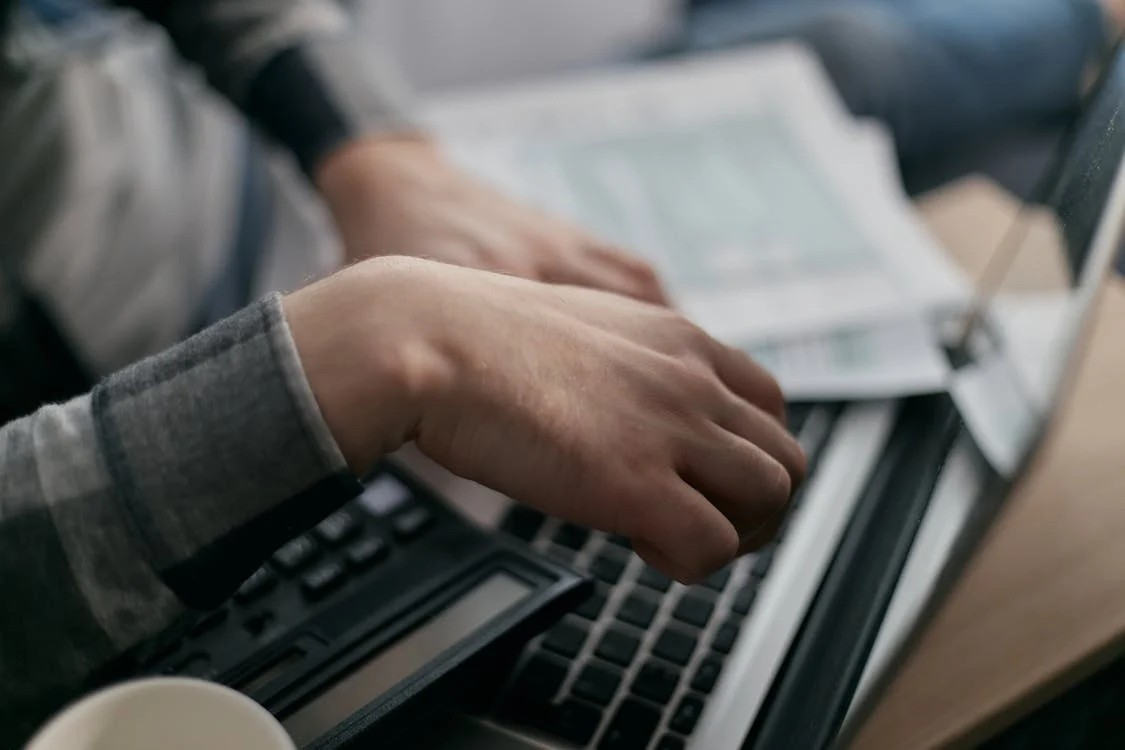 Close-up of hands typing on a laptop and using a calculator, surrounded by financial documents.