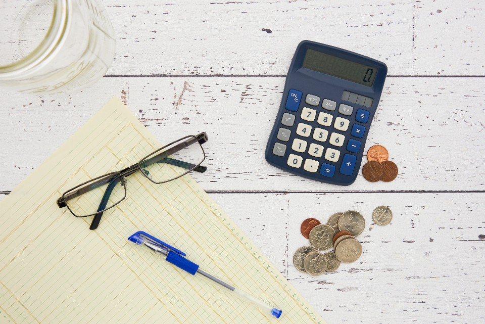 A calculator, coins, a pen, glasses, and a ledger on a table, representing the tools and resources used for managing and calculating credit card debt.