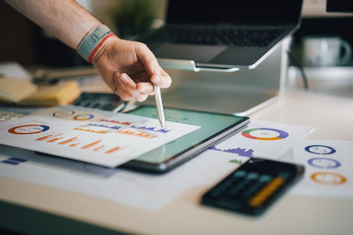 A person pointing at charts and graphs on a tablet, surrounded by financial documents and a calculator.