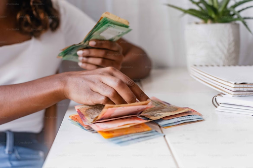 A person counting a stack of various currency notes at a desk with a notebook and a plant in the background.