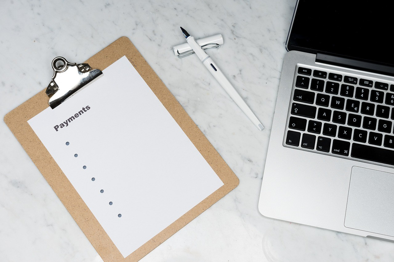 Clipboard with a payments checklist next to a laptop and pen on a marble surface.