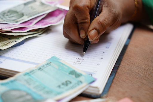 Close-up of a hand writing in a ledger with banknotes on the table, representing the meticulous tracking and planning necessary for achieving debt relief.