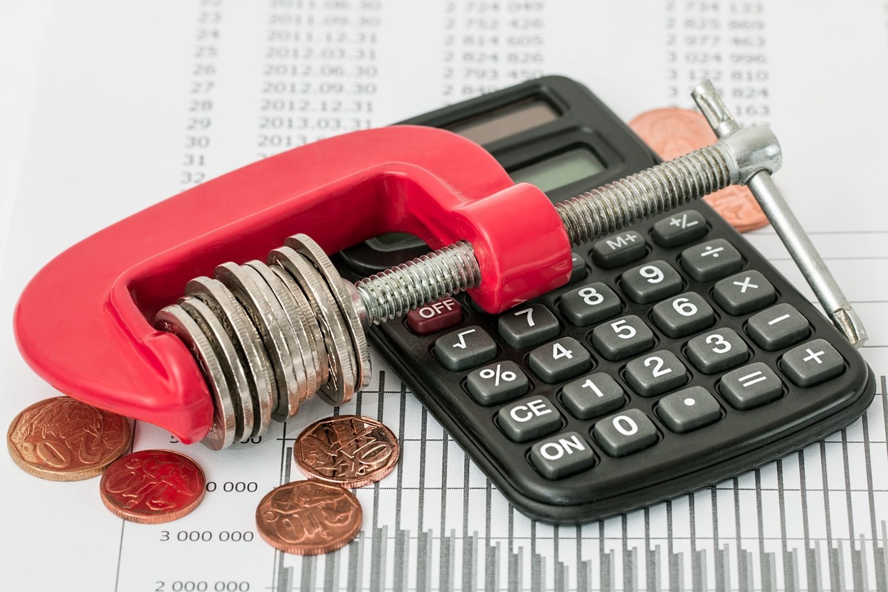 A clamp squeezing coins next to a calculator, symbolizing financial pressure