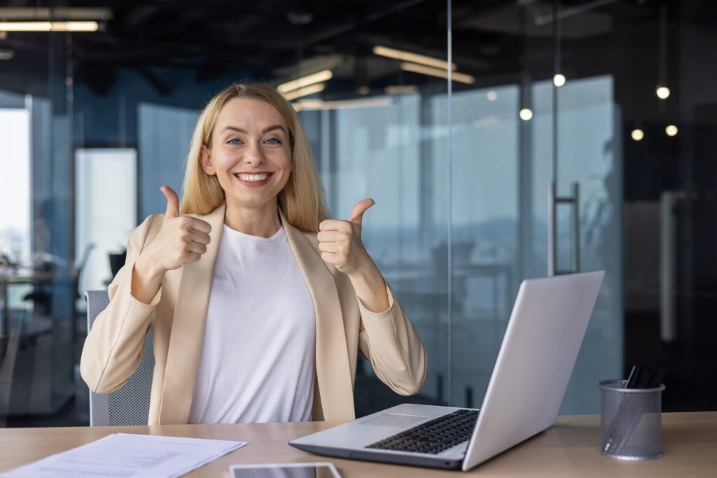 Portrait of a young happy successful businesswoman sitting in the office at the table, smiling at the camera and showing the super gesture with her fingers.
